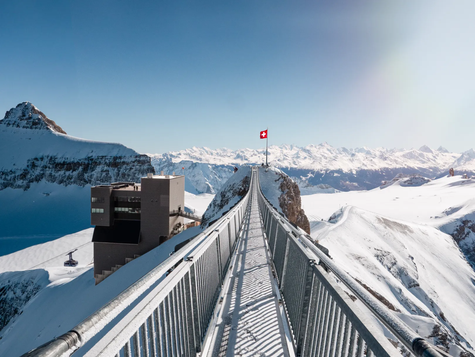 Le Peak Walk by Tissot sur le Glacier 3000 mène au point de vue avec le drapeau suisse, entouré des Alpes enneigées et d’un ciel bleu.