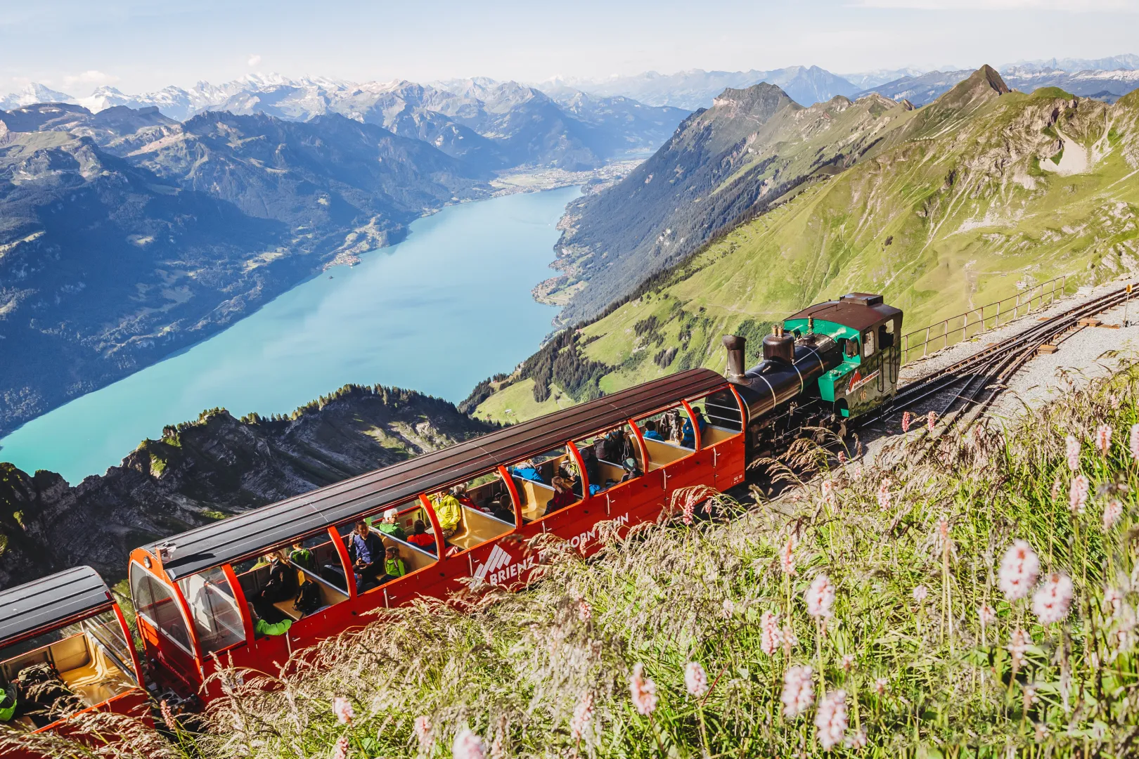 Le chemin de fer du Brienzer Rothorn monte la pente de la montagne avec des wagons rouges ouverts, avec le lac de Brienz turquoise sous un ciel dégagé en arrière-plan.