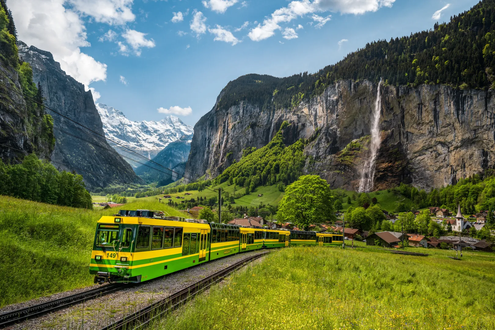 Le Chemin de fer Wengernalp traverse un paysage de prairies verdoyantes à Lauterbrunnen, avec des falaises escarpées, une haute cascade et des montagnes enneigées en arrière-plan.