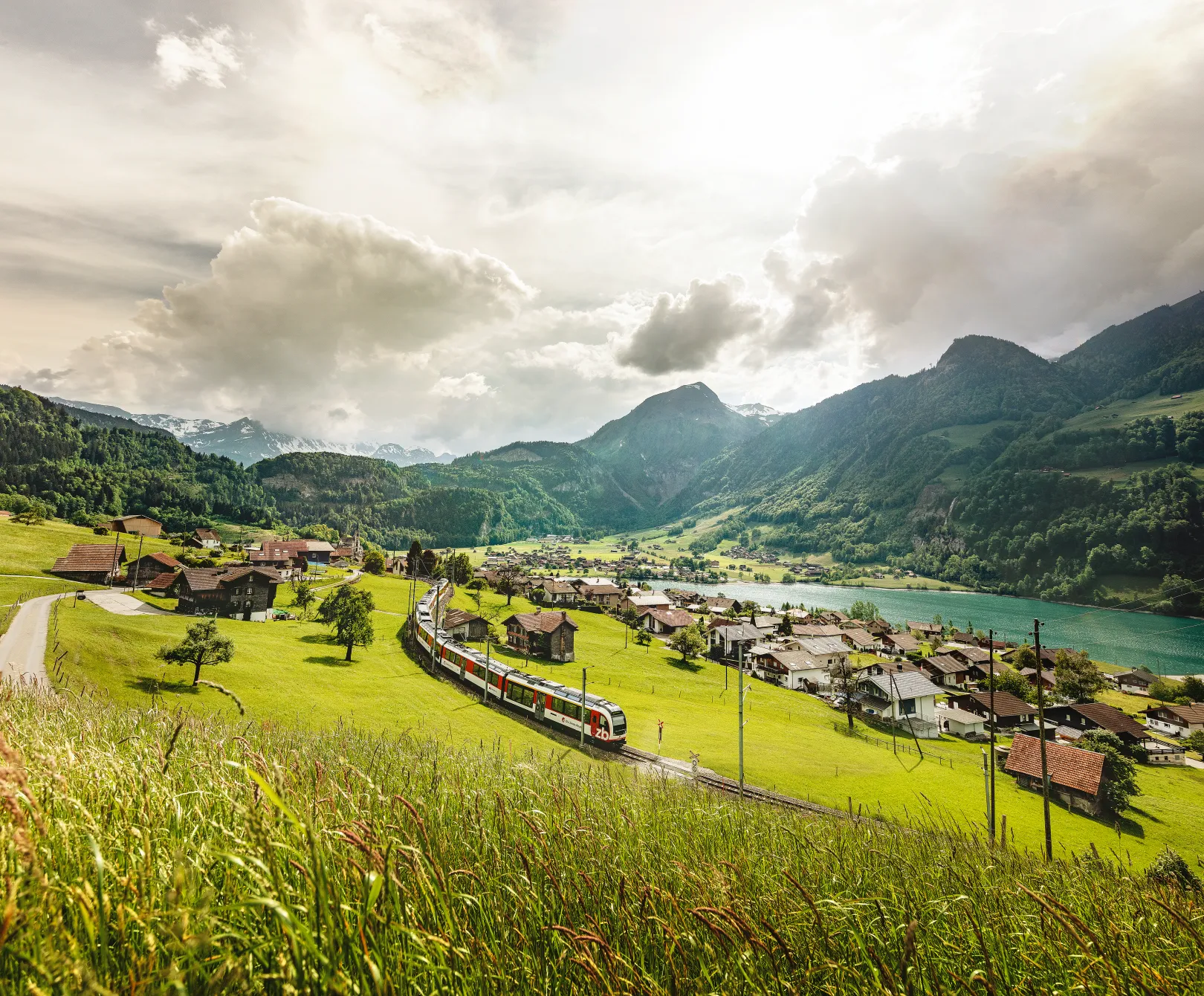 Le Luzern–Interlaken Express traverse en été un paysage montagneux verdoyant à Lungern, avec vue sur un village et un lac.